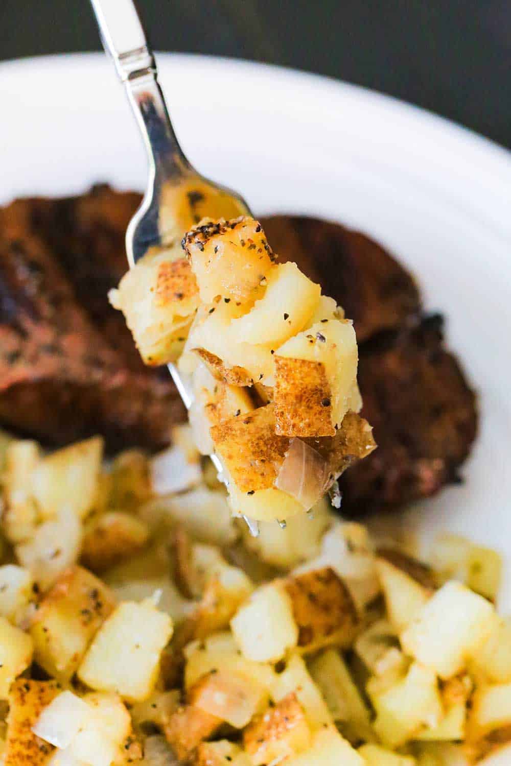 A fork holding up a helping of roasted potatoes in foil over a plate with a grilled steak on it. 