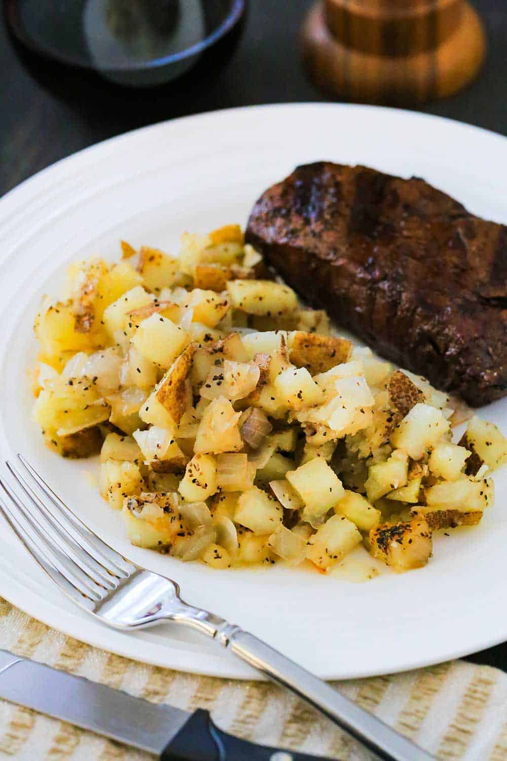 A white dinner plate loaded with roasted potatoes and a grilled steak with a fork nearby. 