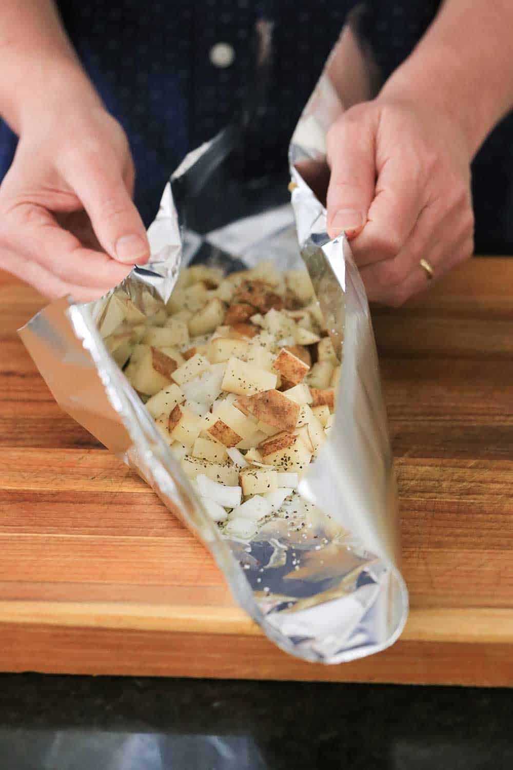 Two hands folding up the sides of a piece of foil over cubed potatoes and chopped onion. 