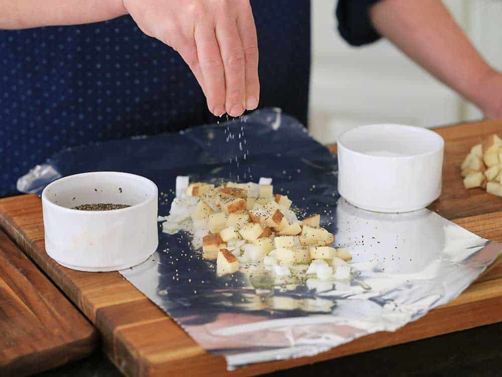 A hand sprinkling salt over cubed potatoes and chopped onions on a piece of foil. 