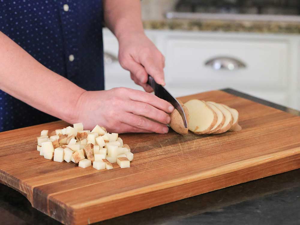Two hands using a chef's knife to slice and cube a russet potato on a large wooden cutting board. 
