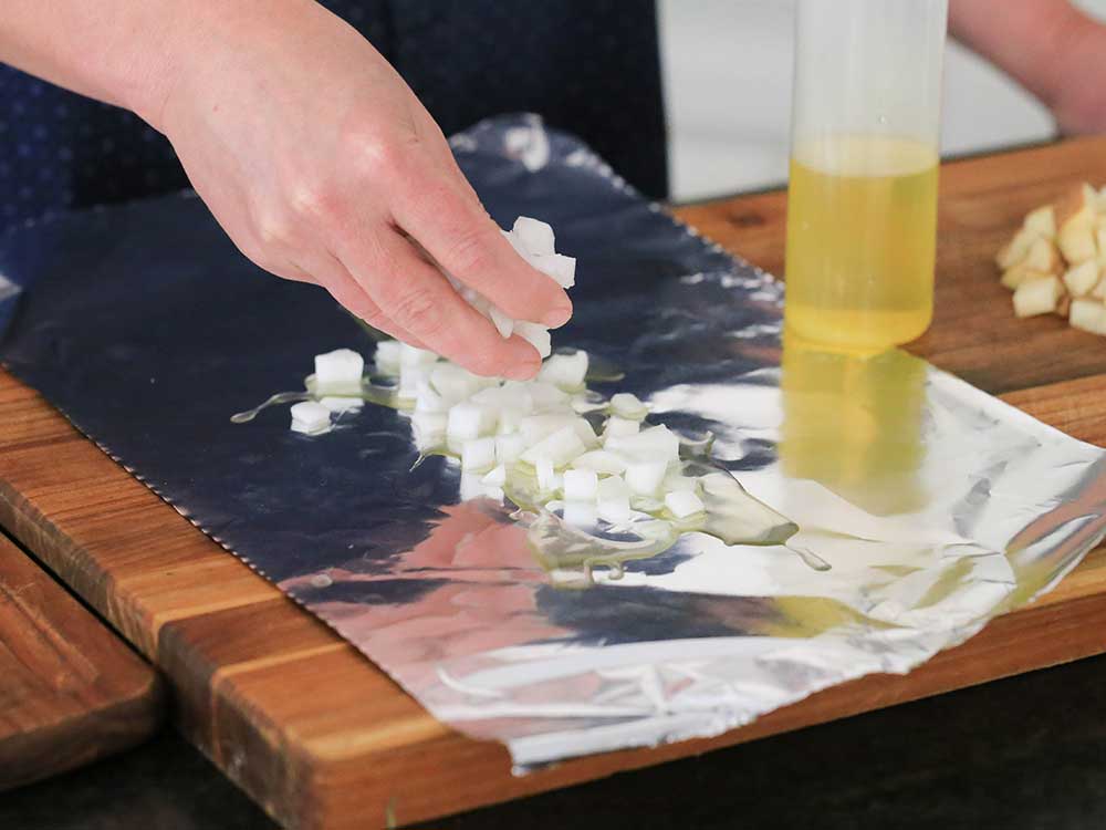 Two hands placing chopped onions on a rectangle of foil with a bottle of oil next to it. 