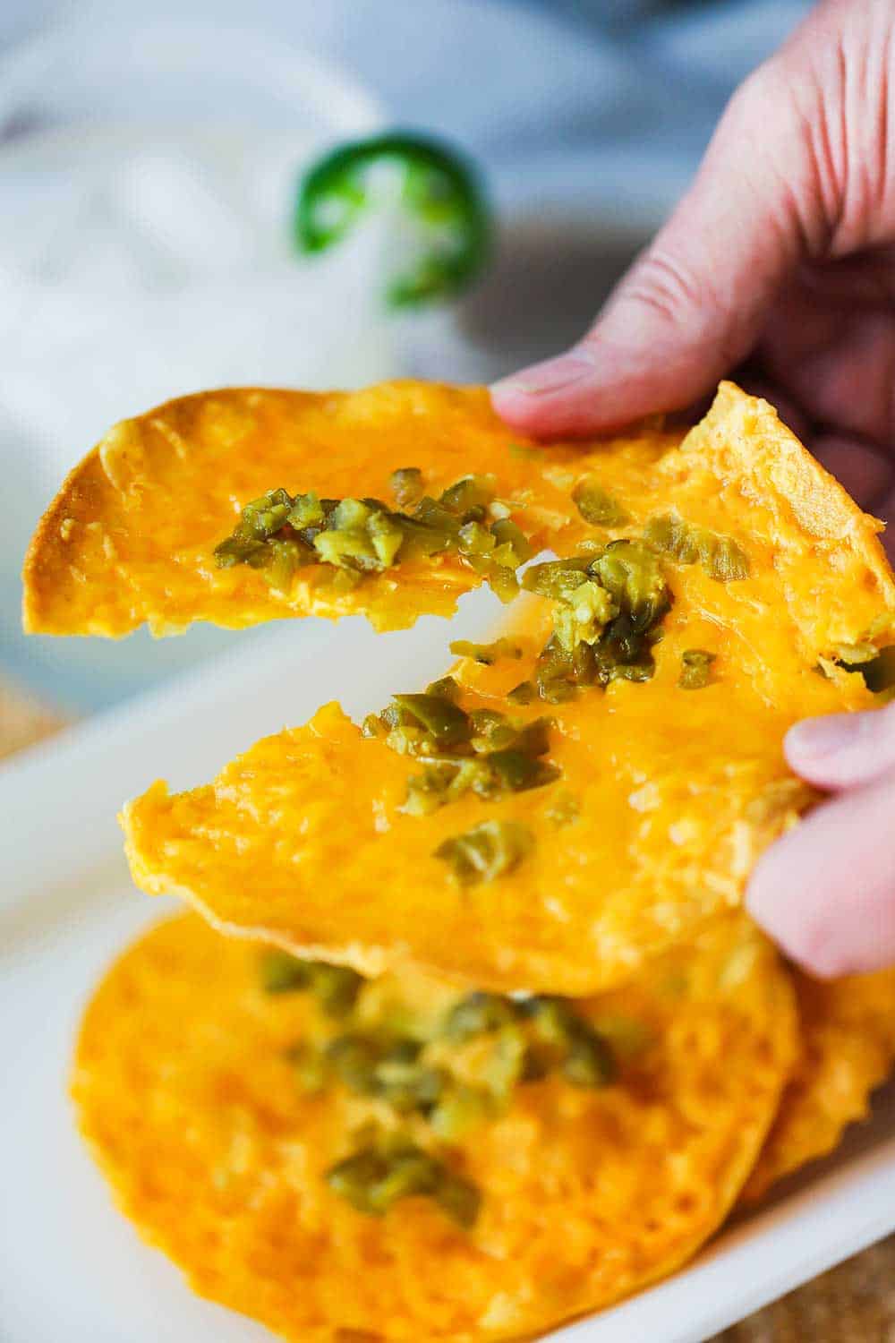 Two hands pulling apart a classic nacho over a platter of them and next to a margarita. 