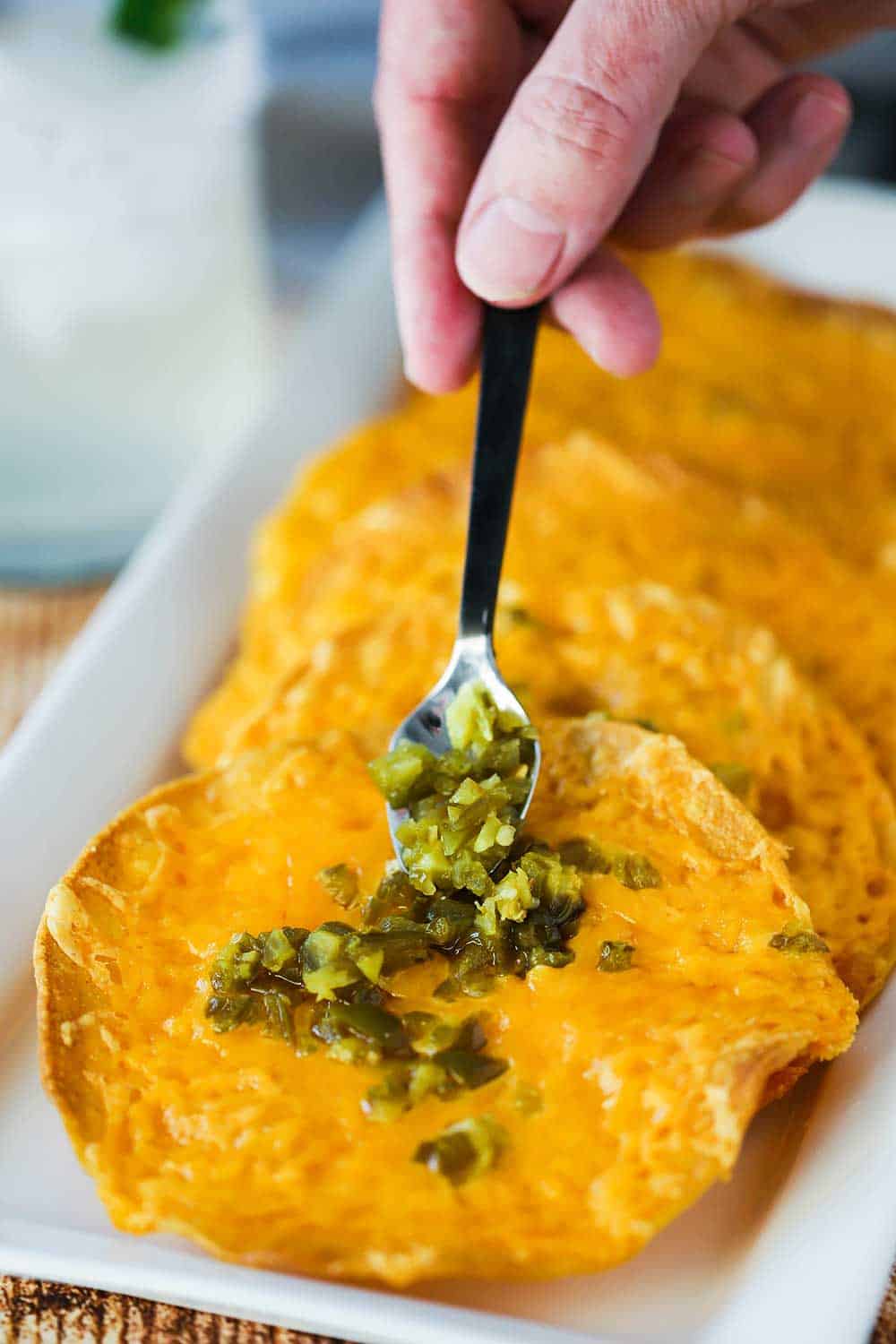 A hand using a small spoon to add diced jalapeños to a stack of classic nachos. 