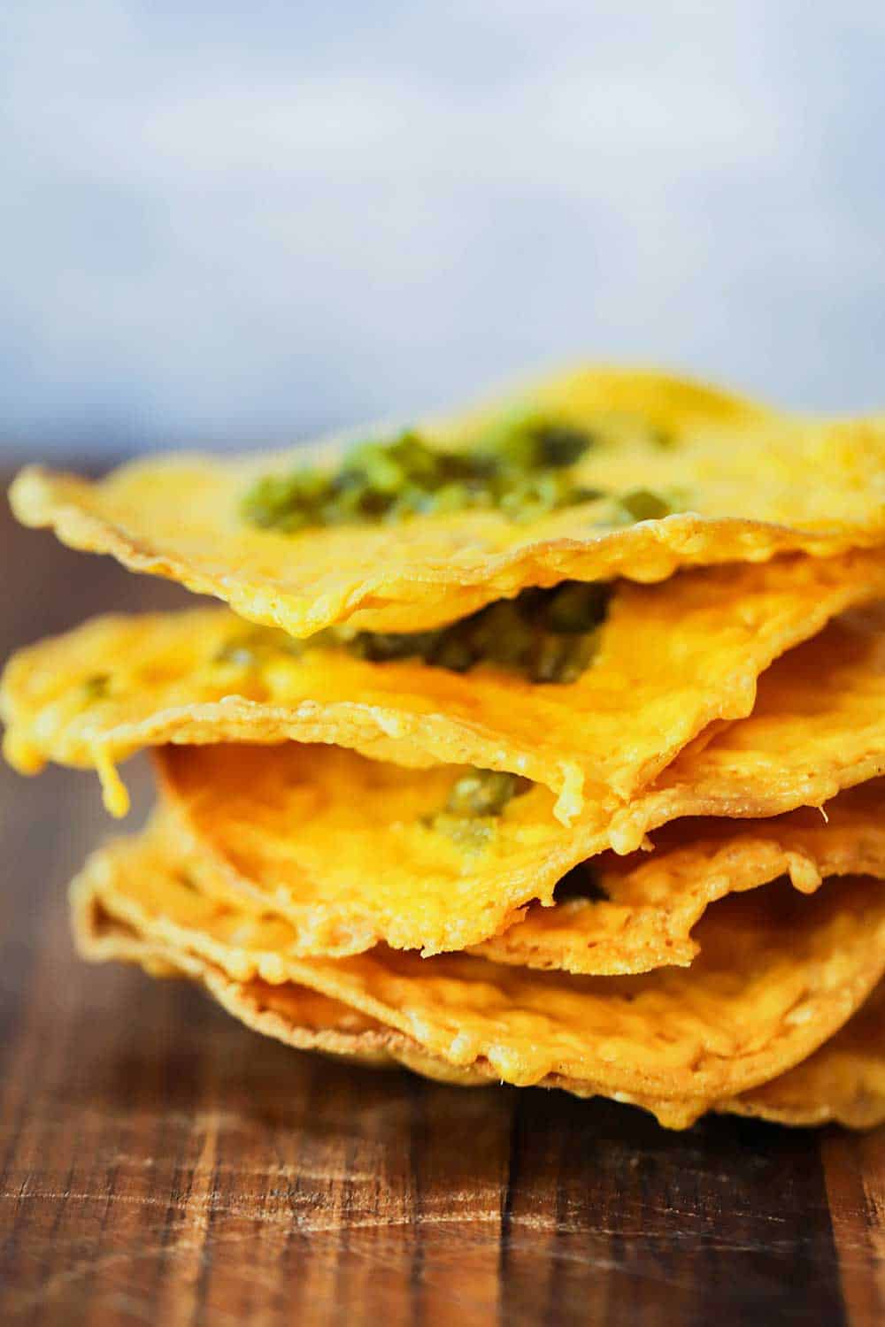 A stack of thin classic nachos topped with jalapeno relish on a wooden cutting board. 