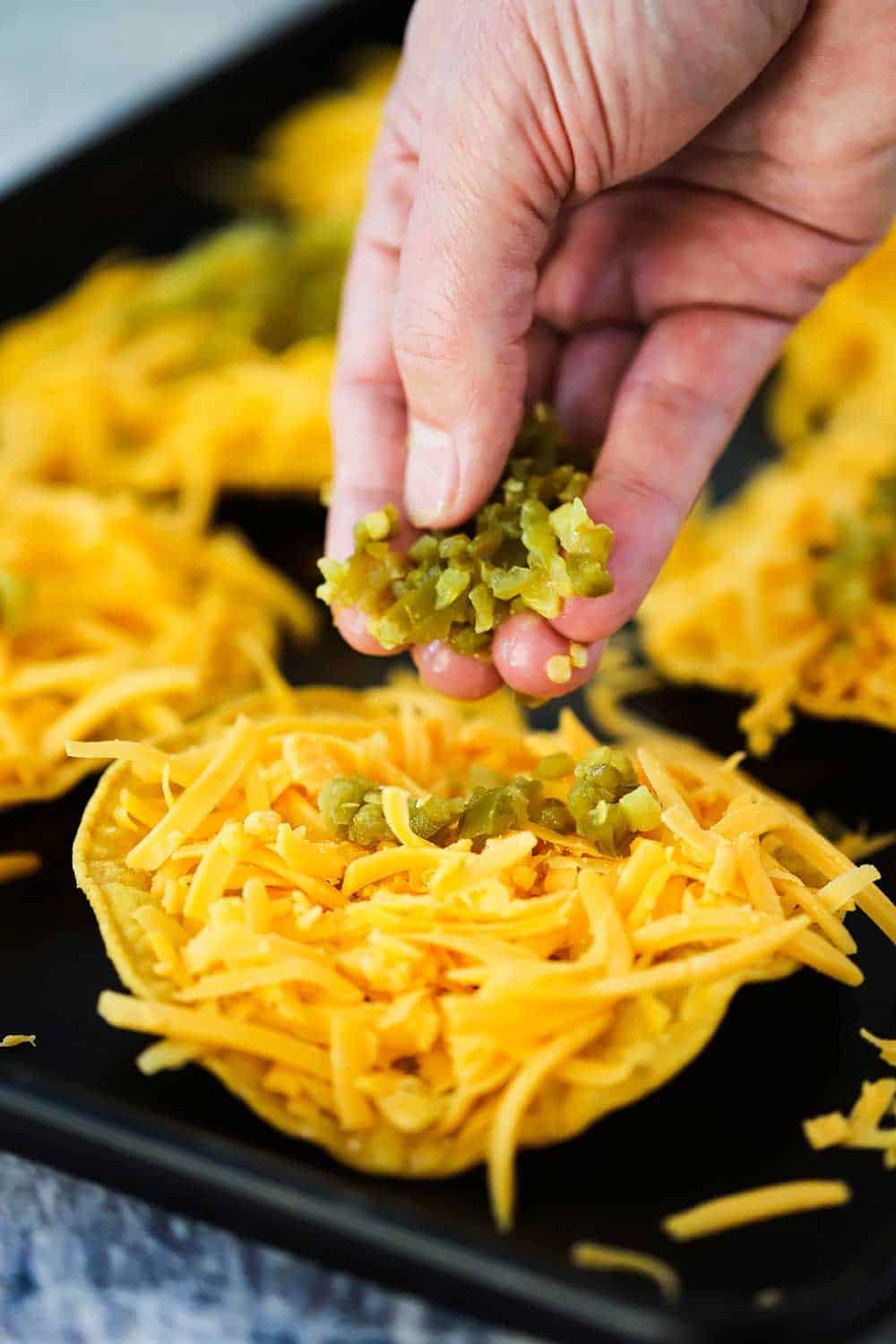 A hand dropping diced jalapenos onto a fried tortilla topped with cheddar cheese on a baking sheet. 