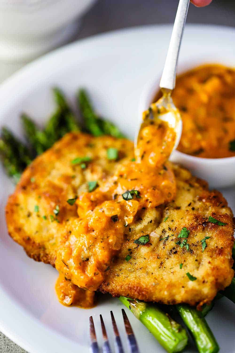 A white plate filled with a layer of roasted asparagus, topped with Parmesan crusted chicken and a spoon adding a layer of vodka sauce on top.