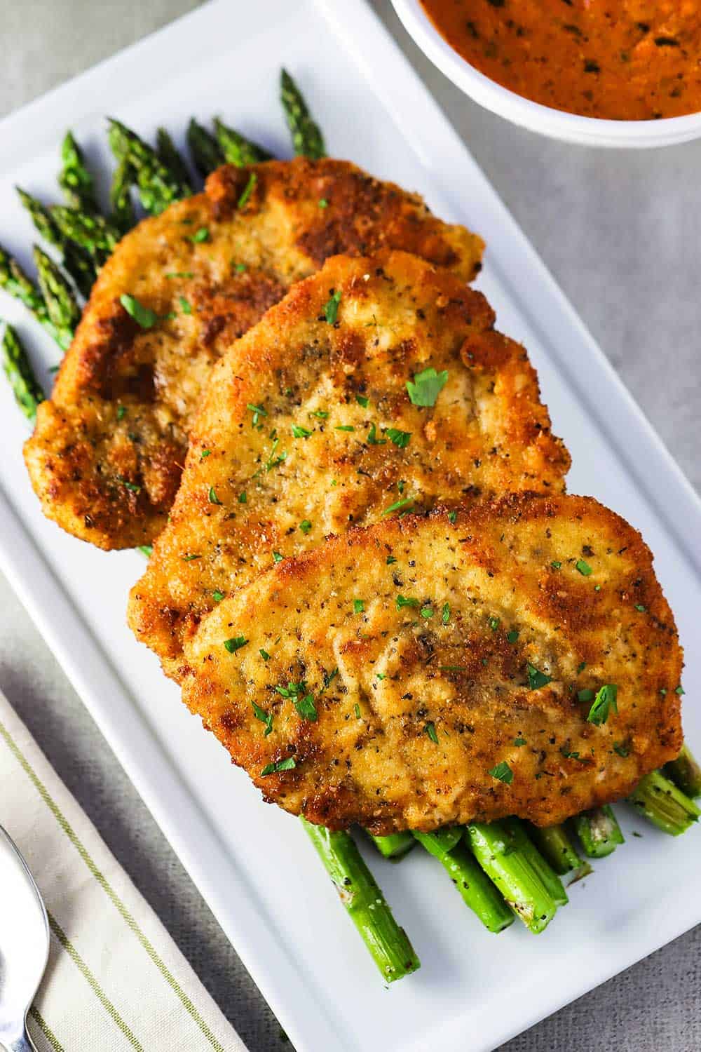 A white serving platter of roasted asparagus with three Parmesan crusted chicken cutlets on top. 