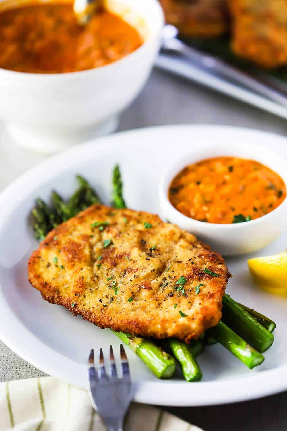 A white dinner plate filled with roasted asparagus topped with Parmesan crusted chicken with a small bowl of vodka sauce next to it.