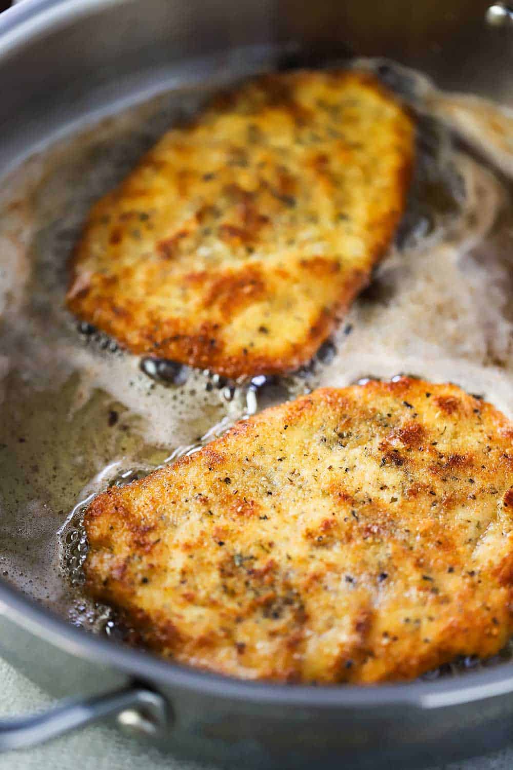 Two Parmesan chicken cutlets cooking in butter in a later steel skillet. 