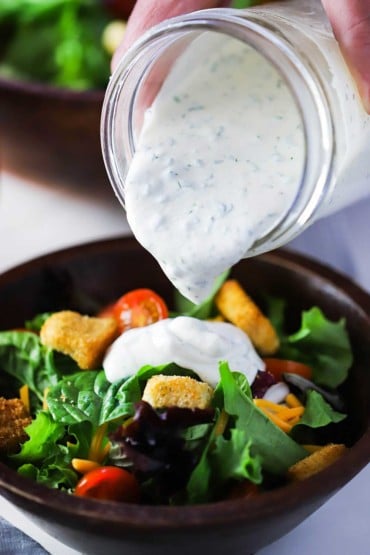 A hand pouring homemade ranch dressing onto a green salad in a brown wooden bowl.