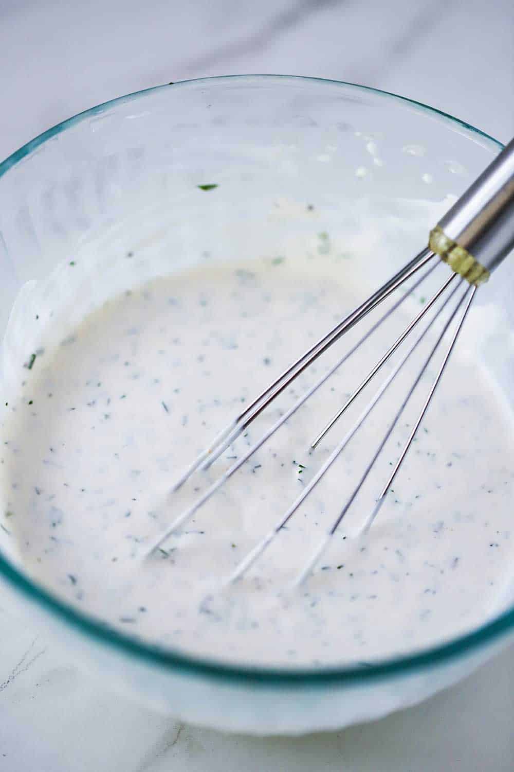 A glass bowl filled with ranch dressing with a wire whisk in it. 