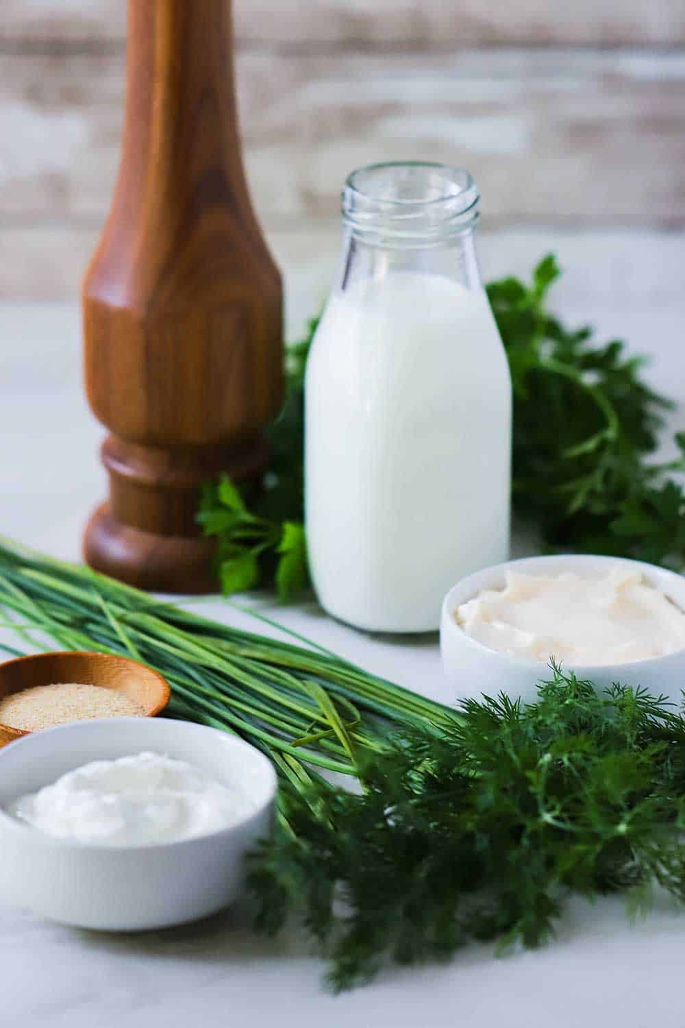 A layout of the ingredients for homemade ranch dressing including bowls of sour cream, mayo, buttermilk, and fresh herbs and a pepper grinder. 