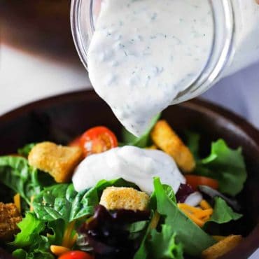 A hand pouring homemade ranch dressing onto a green salad in a brown wooden bowl.