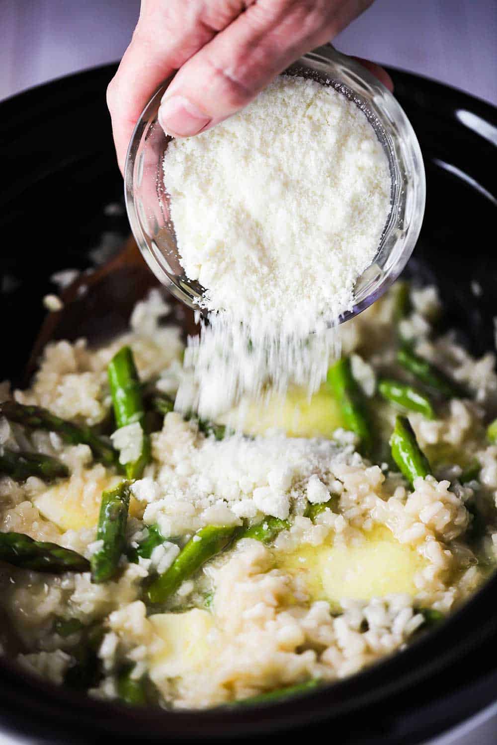 A hand dumping grated Parmesan cheese from a small bowl into a slow cooker filled with asparagus risotto. 