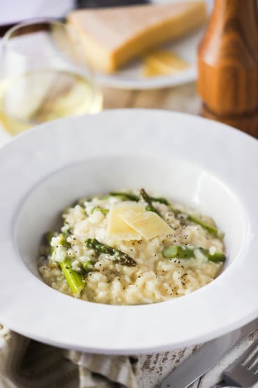 A large white bowl filled with slow cooker asparagus risotto next to a pepper grinder and a glass of white wine.
