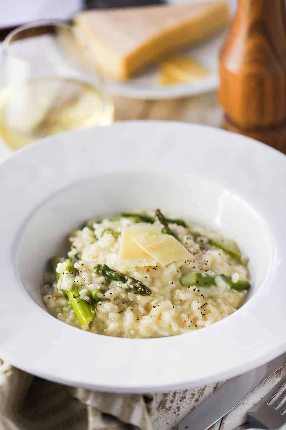 A large white bowl filled with slow cooker asparagus risotto next to a pepper grinder and a glass of white wine. 