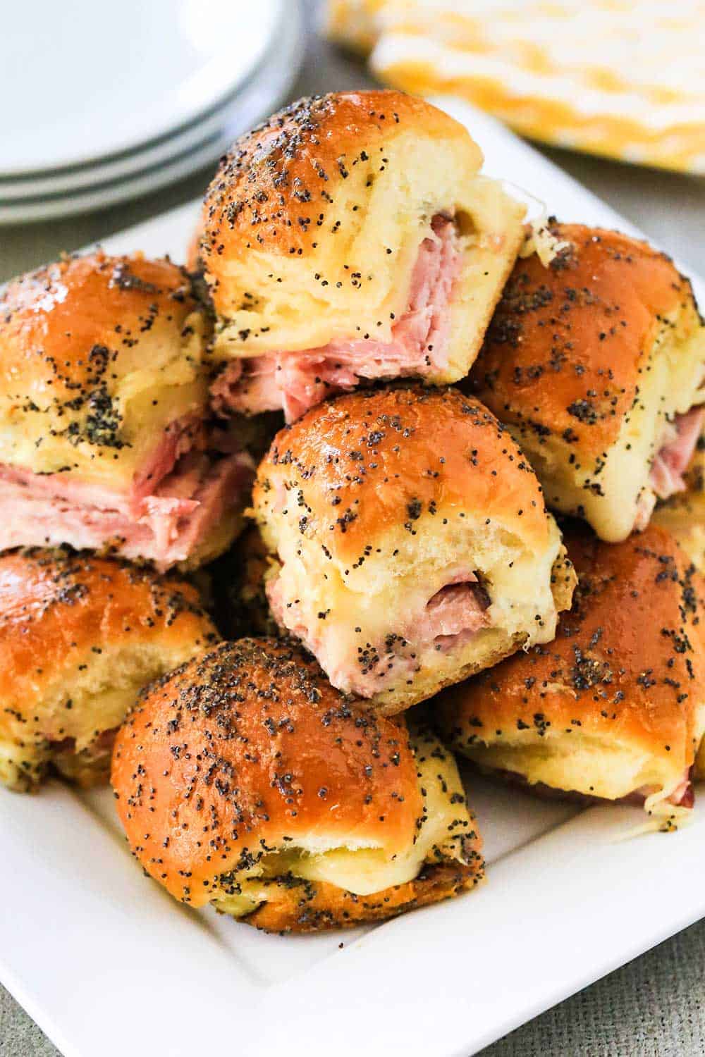 A white plate containing a pile of ham and Swiss sliders next to a yellow napkin. 