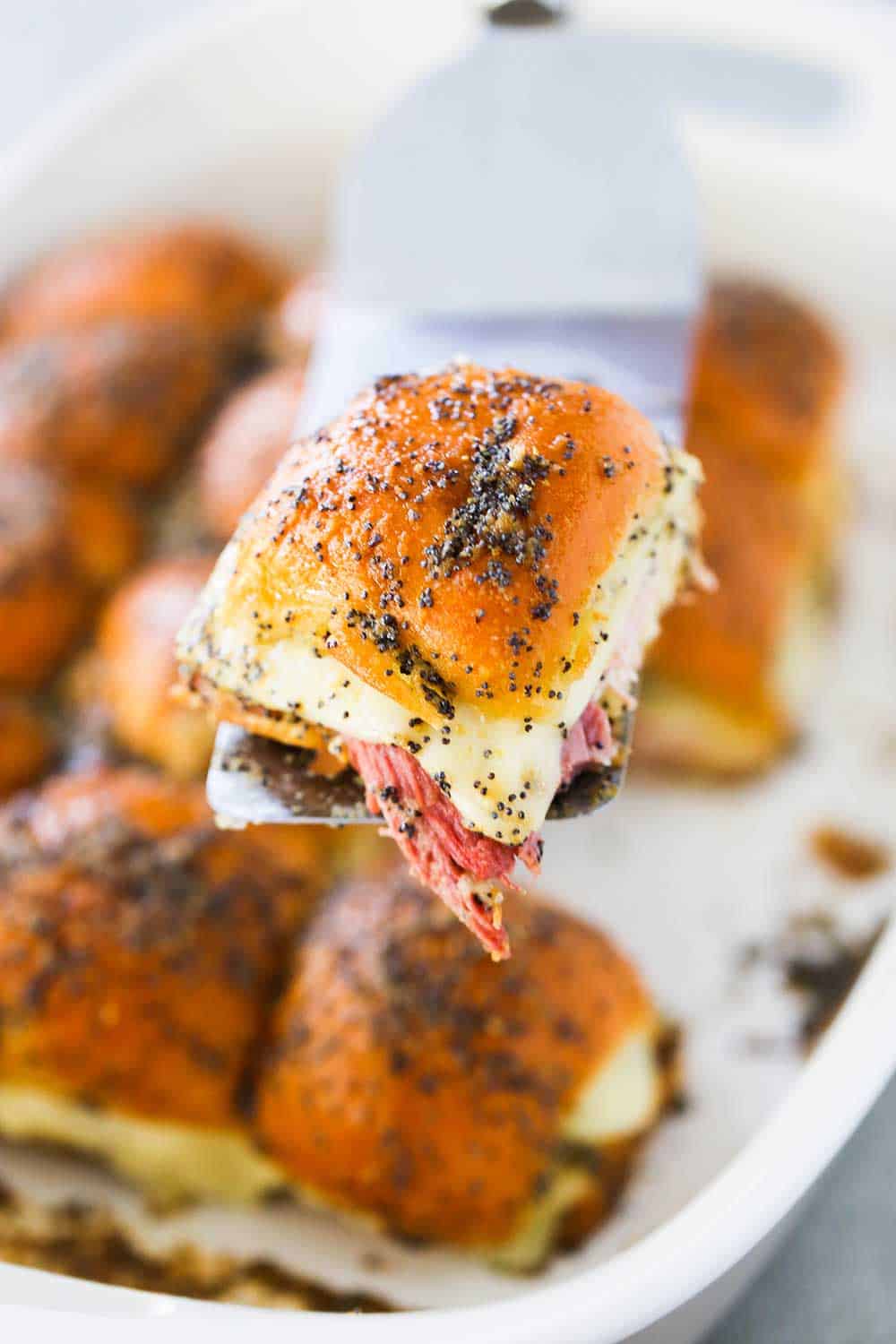 A spatula holding up a ham and Swiss slider over a baking dish filled with them.