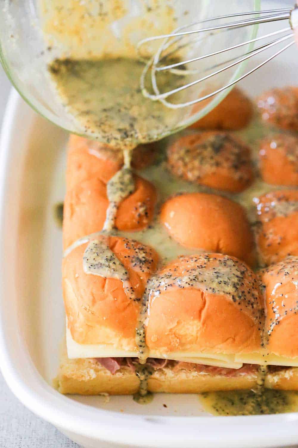 A hand pouring a butter sauce over the tops of ham and Swiss sliders in a white baking dish. 