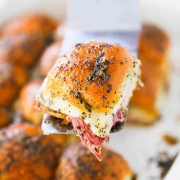 A spatula holding up a ham and Swiss slider over a baking dish filled with them.