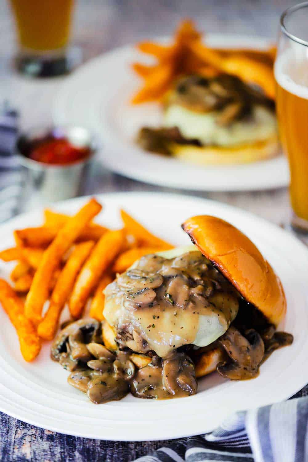 A mushroom Swiss burger on a white plate next to a small bowl of ketchup and and a beer. 