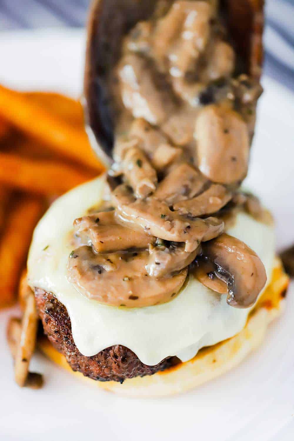 A large wooden spoon placing mushroom sauce onto a mushroom Swiss burger on a plate next to a pile of French fries. 