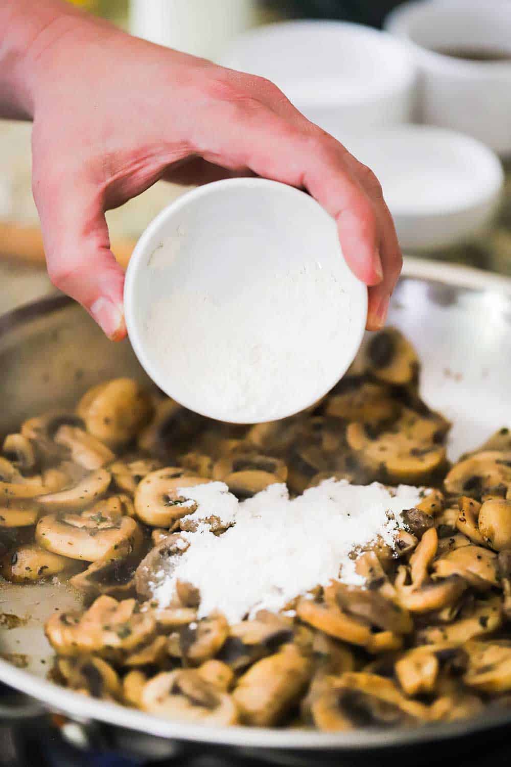 A hand sprinkling flour from a small bowl into a skillet filled with sautéed mushrooms.