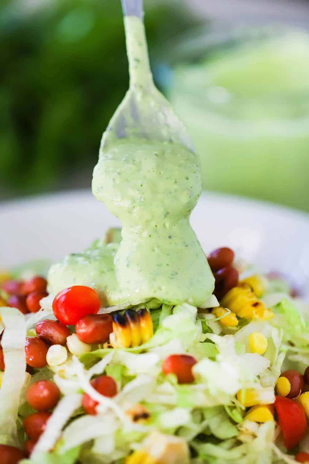 A spoon drizzling avocado lime over the top of a chopped salad in a white bowl topped with grilled corn, cherry tomatoes, and pinto beans. 