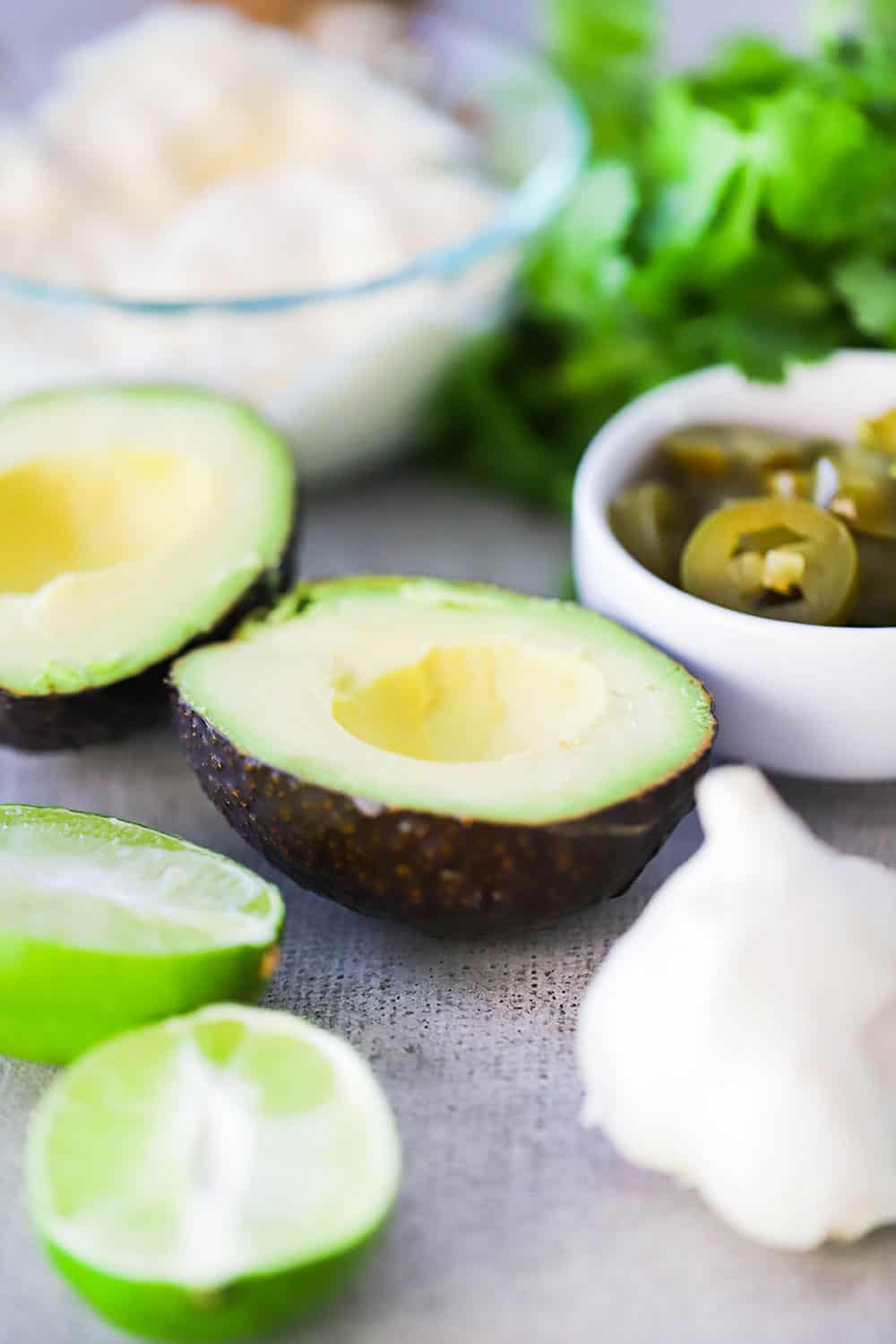 Ingredients for avocado lime dressing on a grey board including a cut open avocado, cut limes, a head of garlic, fresh cilantro, and a small bowl of mayonnaise. 