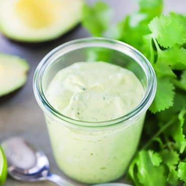 A small glass jar holding freshly made avocado lime dressing next to a small spoon and a bunch of fresh parsley and a cut avocado in the background.