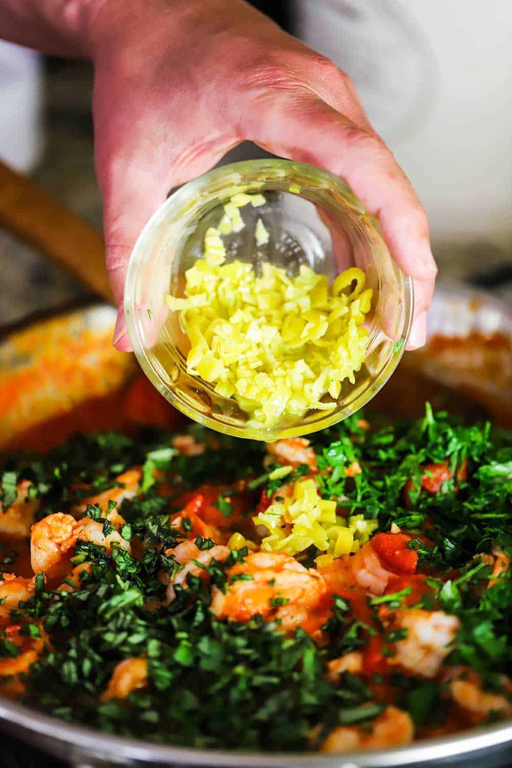 A hand dumping chopped pepperoncini from a small bowl into a skillet filled with cooked shrimp, tomatoes, and fresh herbs.