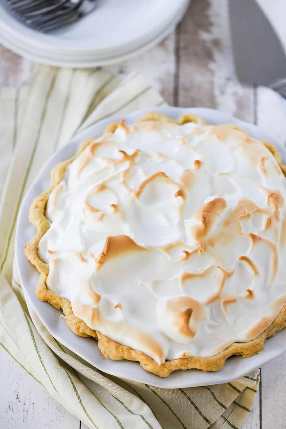 An overhead view of a lemon meringue pie with the topping lightly browned and sitting next to a striped napkin and dessert plates with forks.