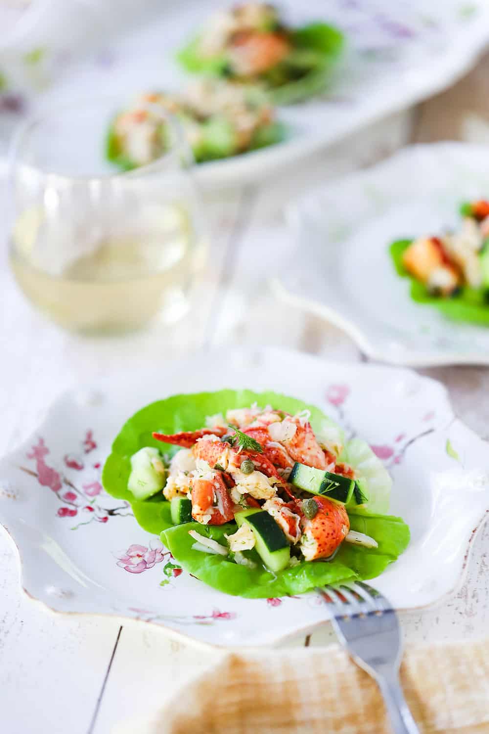 A mound of lobster salad with cucumber on a leaf of lettuce on a decorative plate next to a glass of white wine and a platter.