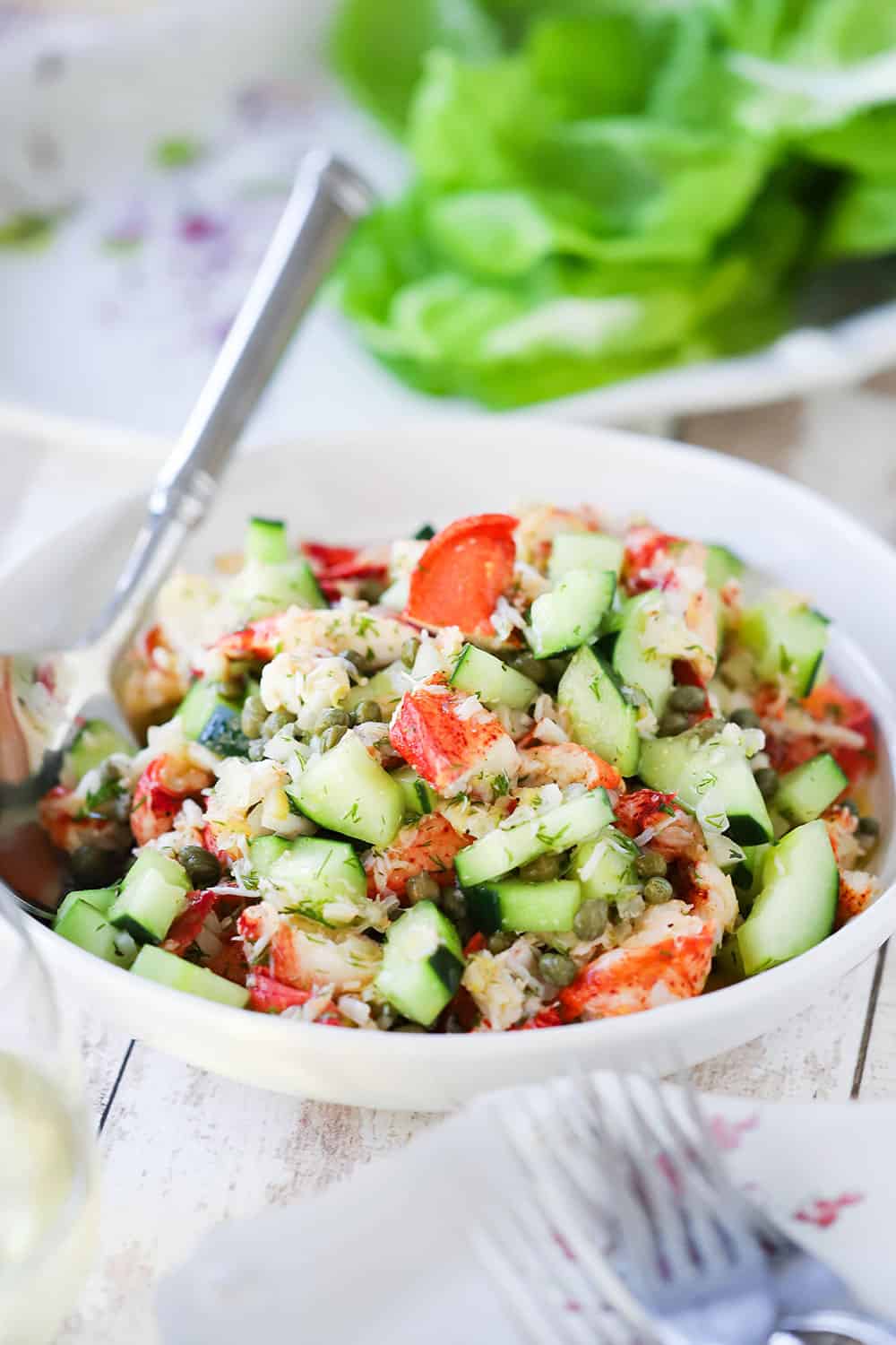 A white serving bowl filled with a lobster salad with cucumber and herbs with a large silver spoon in the salad.