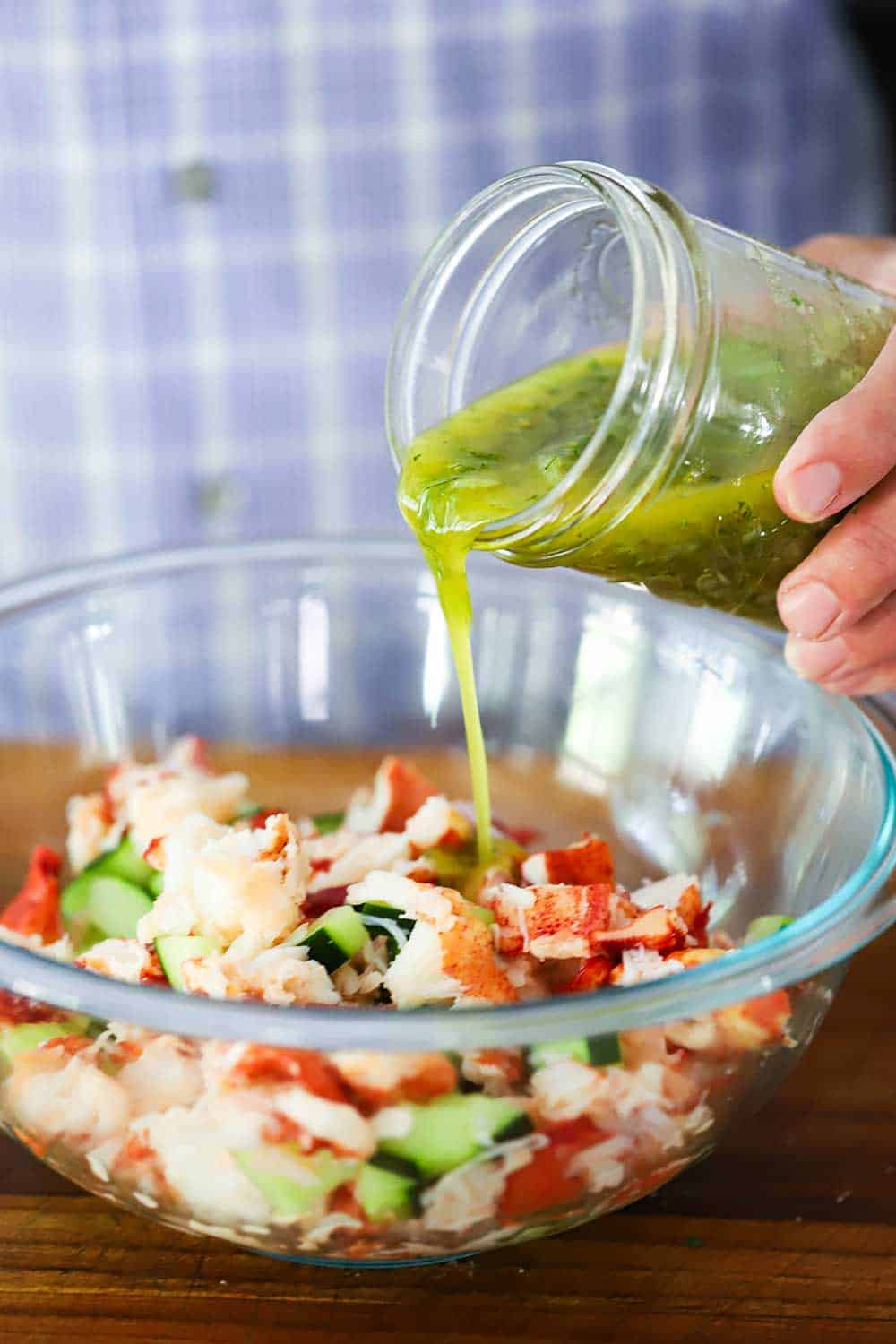 A person pouring a lemon vinaigrette from a mason jar into a glass bowl filled with chopped fresh lobster and cucumbers.