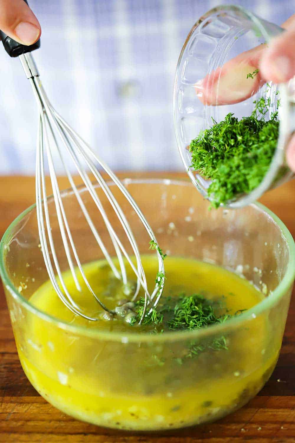 A hand dropping fresh dill from a small bowl into another glass bowl filled with lemon vinaigrette with a whisk in it.