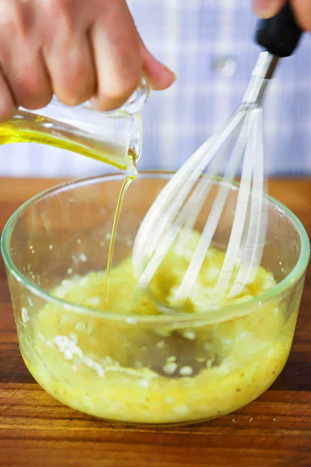 A hand pouring olive oil from a glass vessel into a glass bowl filled with lemon vinaigrette with a whisk in it.