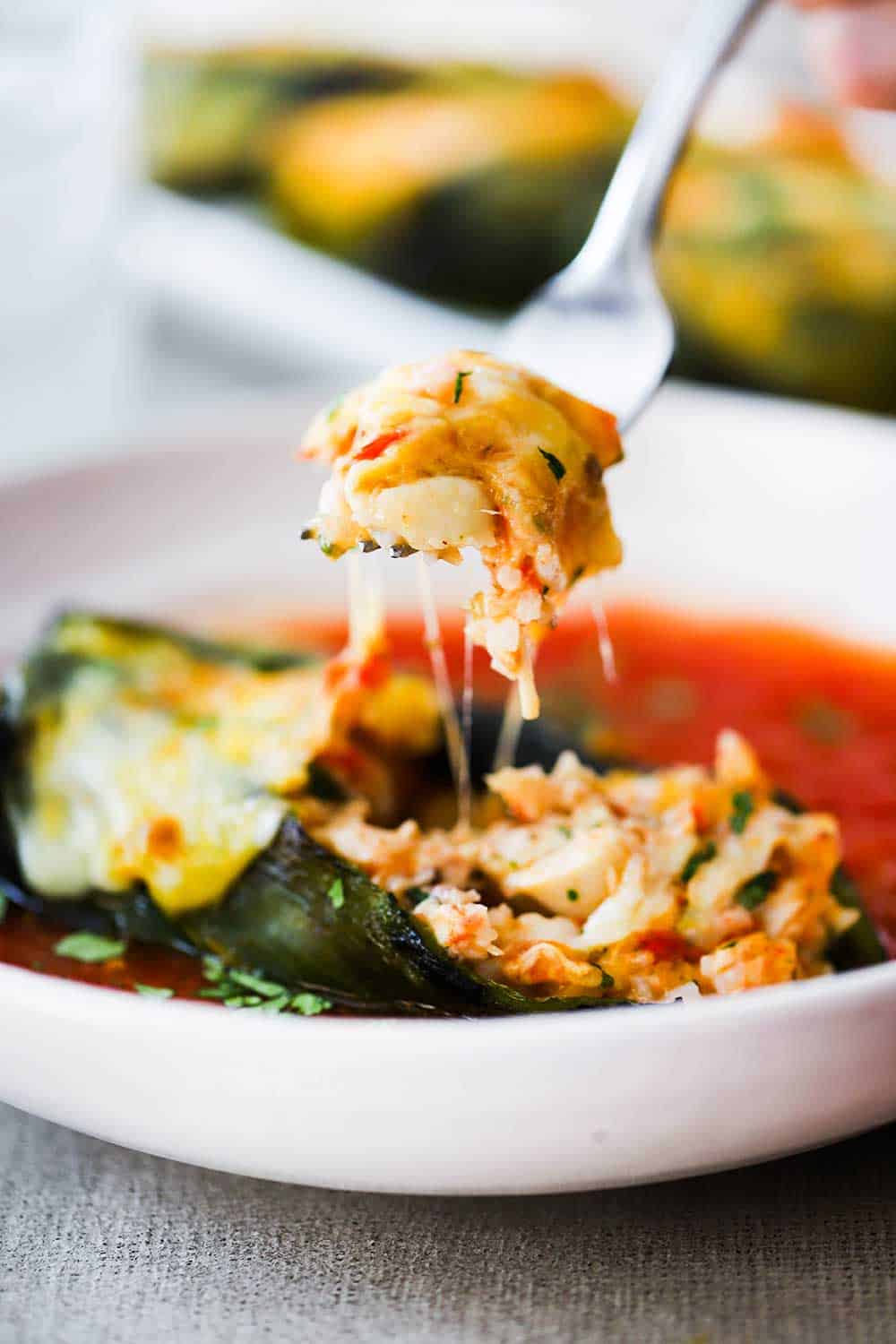 A hand holding a fork filled with a chicken and rice stuffing from inside a roasted poblano in a white bowl with red sauce. 