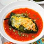 An overhead view of a stuffed poblano pepper topped with melted cheese in a white serving bowl filled with a red sauce.