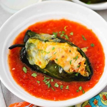 An overhead view of a stuffed poblano pepper topped with melted cheese in a white serving bowl filled with a red sauce.