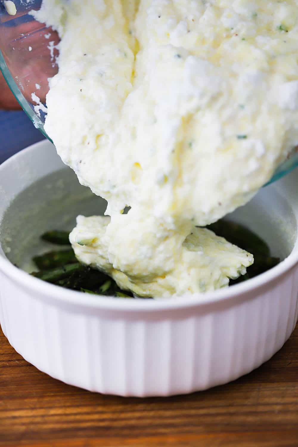 An uncooked souffle mixture being dropped into a white souffle bowl filled with roasted asparagus.