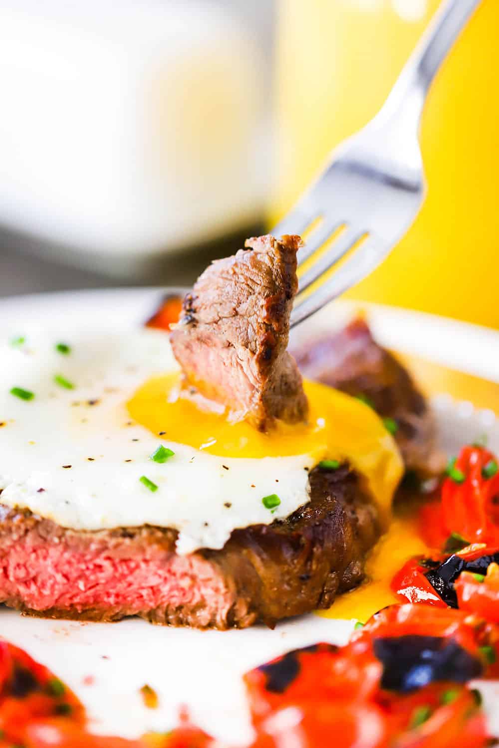 A fork with a piece of steak stuck into it is being plunged into the yolk of an egg sitting on top of a seared steak next to roasted cherry tomatoes.