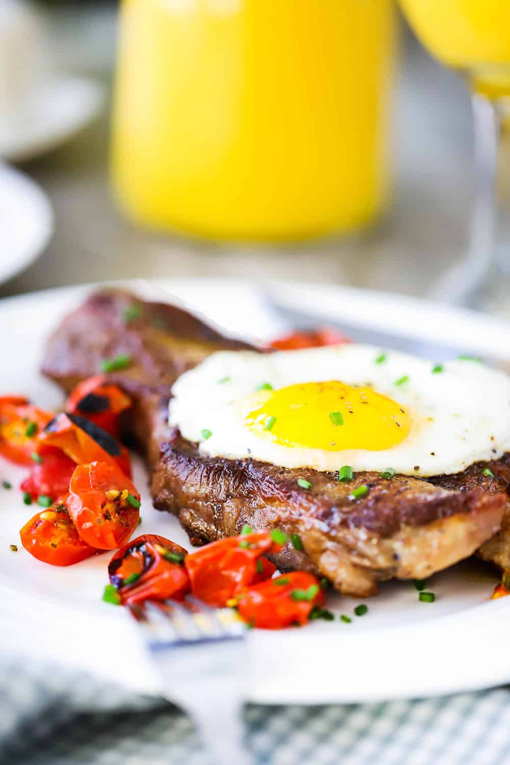 A close up view of a seared New York strip steak on a white plate with a fried egg on top of it next to roasted cherry tomatoes with orange juice in the background.