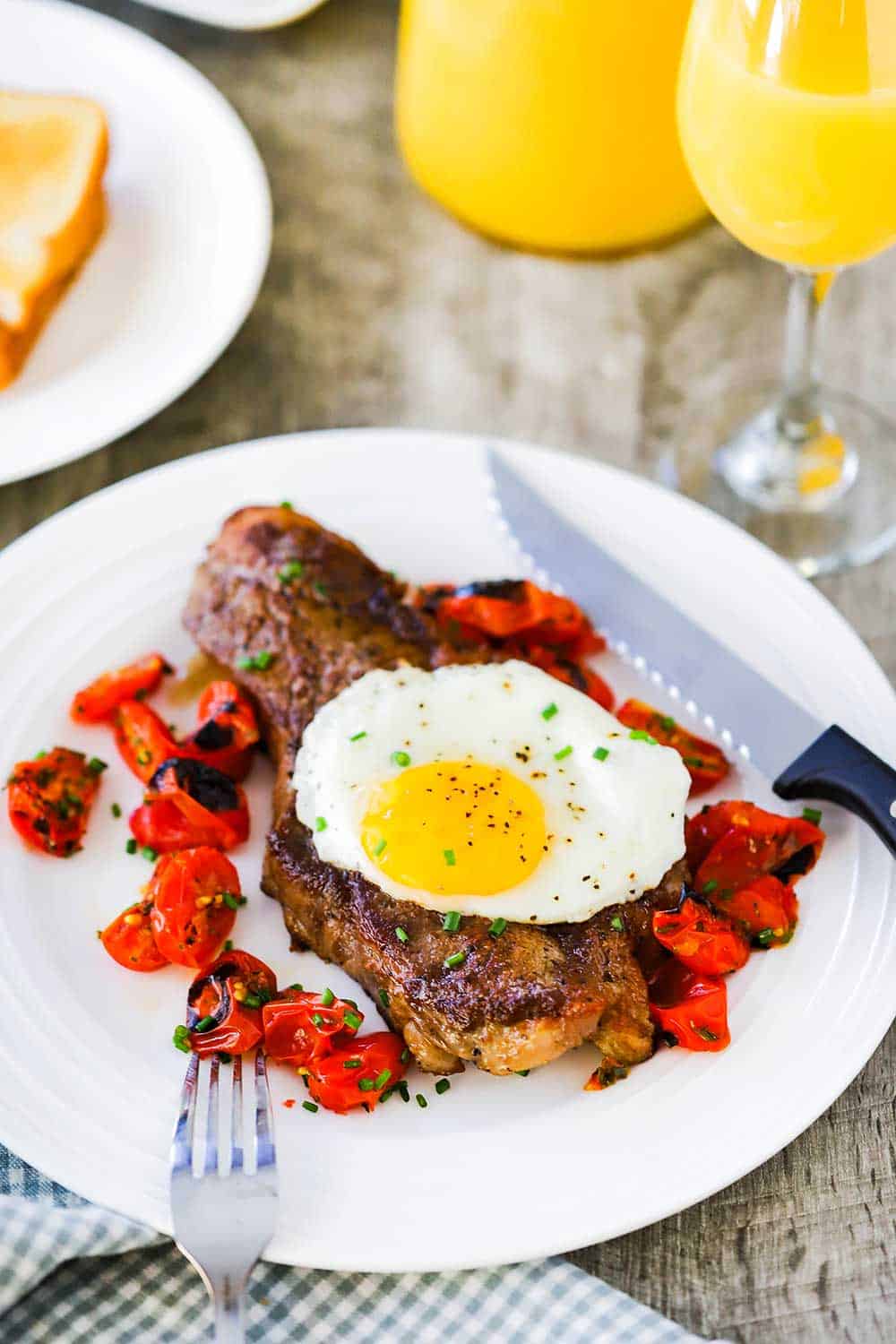A view looking down on a white plate filled with a seared New York strip steak topped with a fried egg next to roasted tomatoes with a glass of orange juice nearby.
