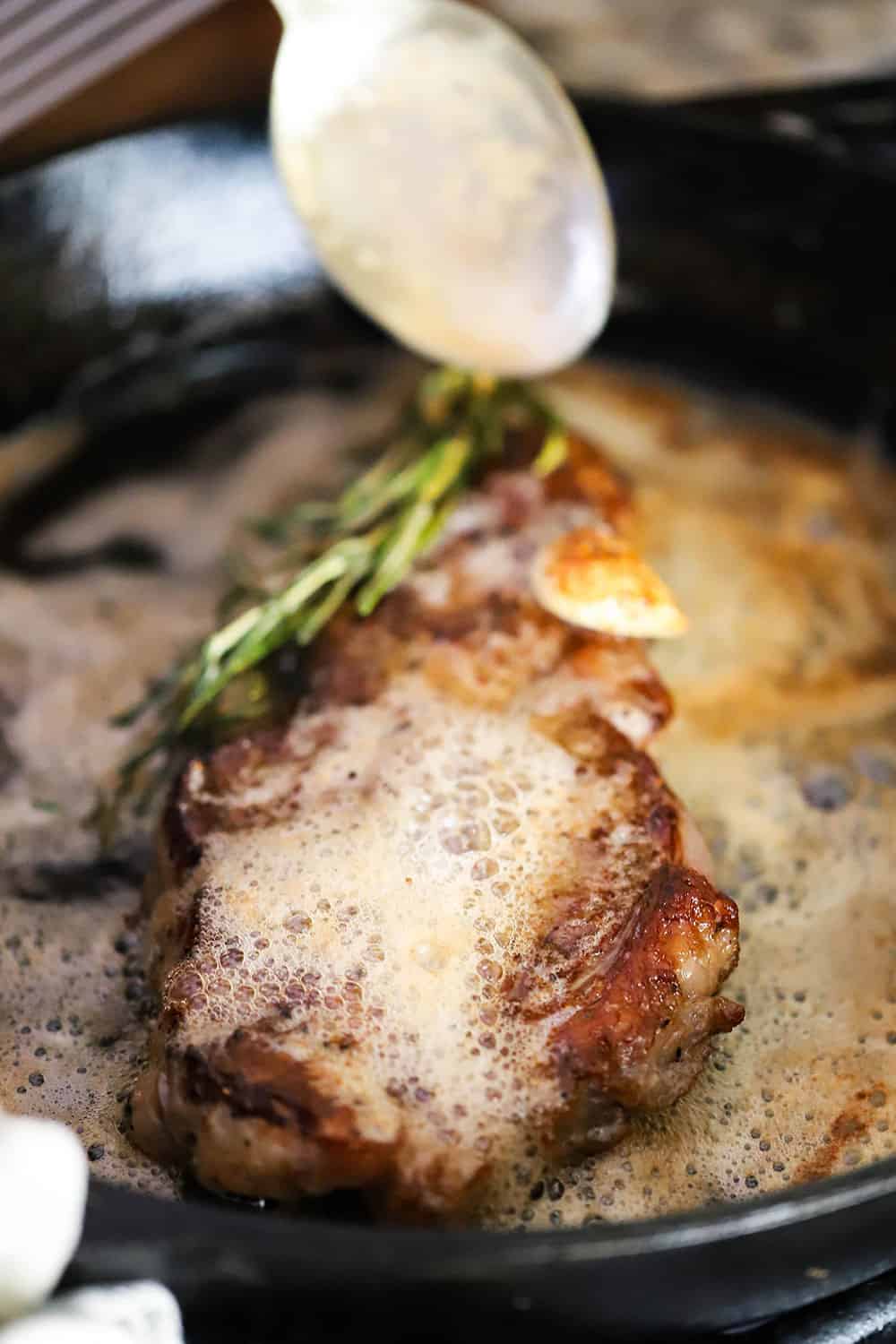 A spoon pouring hot butter over a seared New York strip steak in a cast iron skillet with rosemary and garlic on the steak.