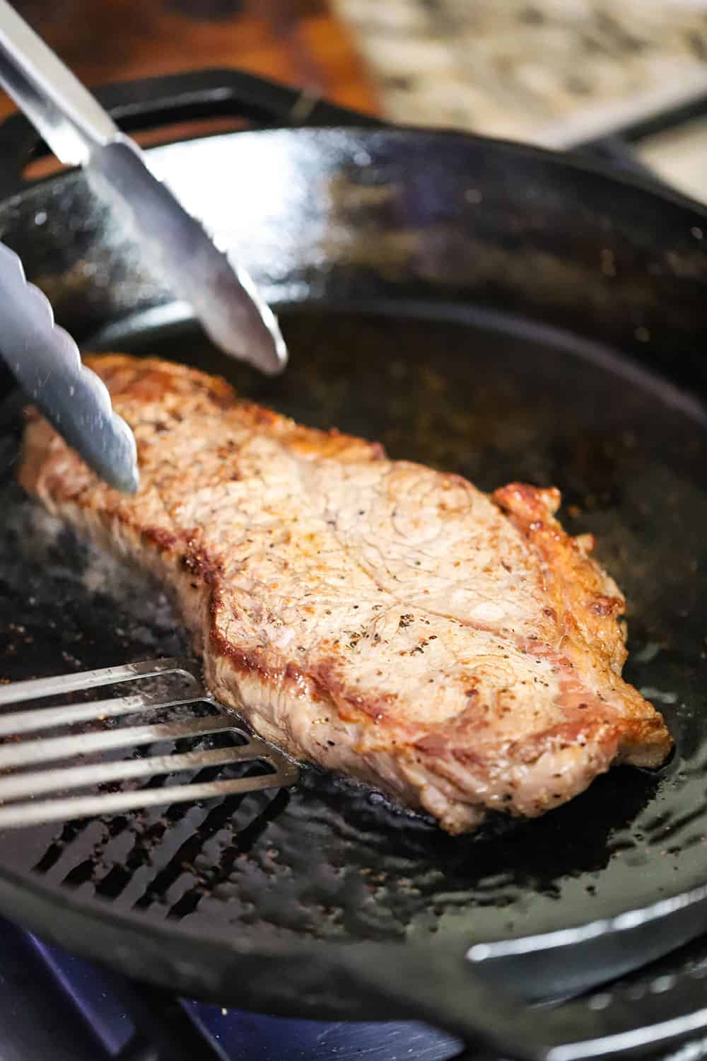 A New York strip steak being seared int a cast iron skillet with a pair of tongs and metal spatula getting ready to flip it over.