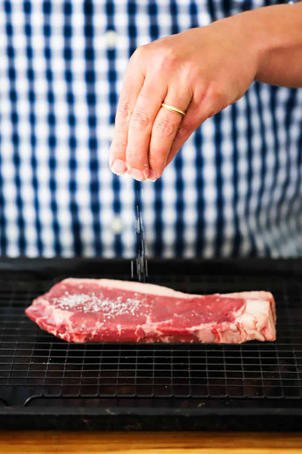 A hand sprinkling salt all over an uncooked New York strip steak on a baking rack fitted into a baking sheet.