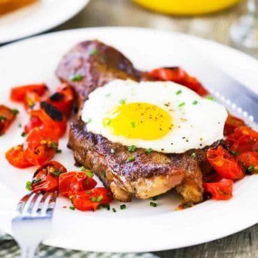 A close up view of a seared New York strip steak on a white plate with a fried egg on top of it next to roasted cherry tomatoes with orange juice in the background.