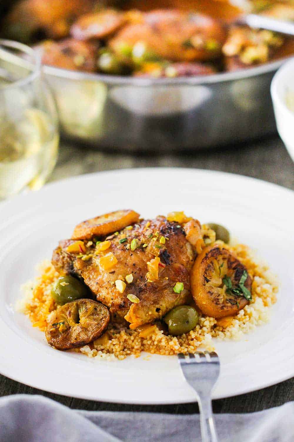 A white dinner plate with a serving of chicken tagine with preserved lemons and olives over a bed of couscous, all sitting next to a skillet filled with the chicken tagine. 